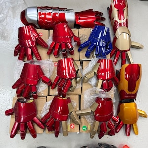 Op de afbeelding: Een verzameling gearticuleerde robotische hand- en armhandschoenen in metallic rode, gouden en blauwe afwerkingen. De set bevat meerdere individuele handstukken, sommige met gouden accenten, en één volledige armconstructie met een zilver en rood gesegmenteerd ontwerp.
