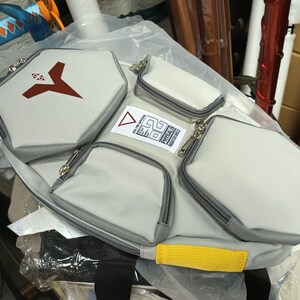 May include: A light gray multi-pocket bag with a black strap and yellow accent. The bag has multiple zippered compartments, including a large hexagonal pocket with a red logo. The bag has a label with text and a triangle.