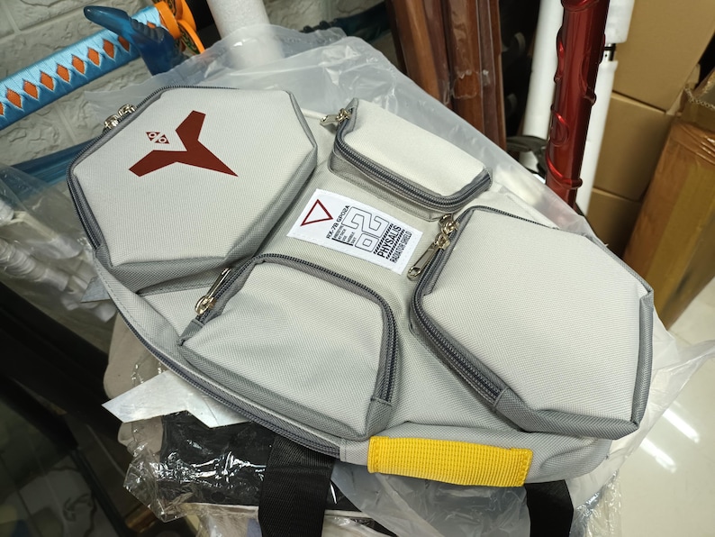 May include: A light gray multi-pocket bag with a black strap and yellow accent. The bag has multiple zippered compartments, including a large hexagonal pocket with a red logo. The bag has a label with text and a triangle.