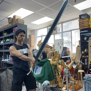 May include: A large, black sword with a silver edge is held by a person in a workshop. The sword has a white handle. Other props and figures are visible in the background, including a gold-colored statue and a Terminator robot.