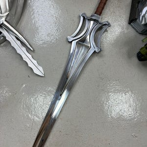 Może przedstawiać: Srebrny miecz fantasy z brązową rękojeścią owiniętą sk&oacute;rą. Ostrze jest długie i wypolerowane, z ozdobnym jelcem z zakrzywionymi elementami. Miecz jest prawdopodobnie rekwizytem lub przedmiotem kolekcjonerskim.