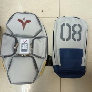May include: Two gray backpacks. One is a unique shape with a red symbol and multiple pockets. The other is rectangular with the number "08" in gray and a blue pocket. Both have black straps and zippers.
