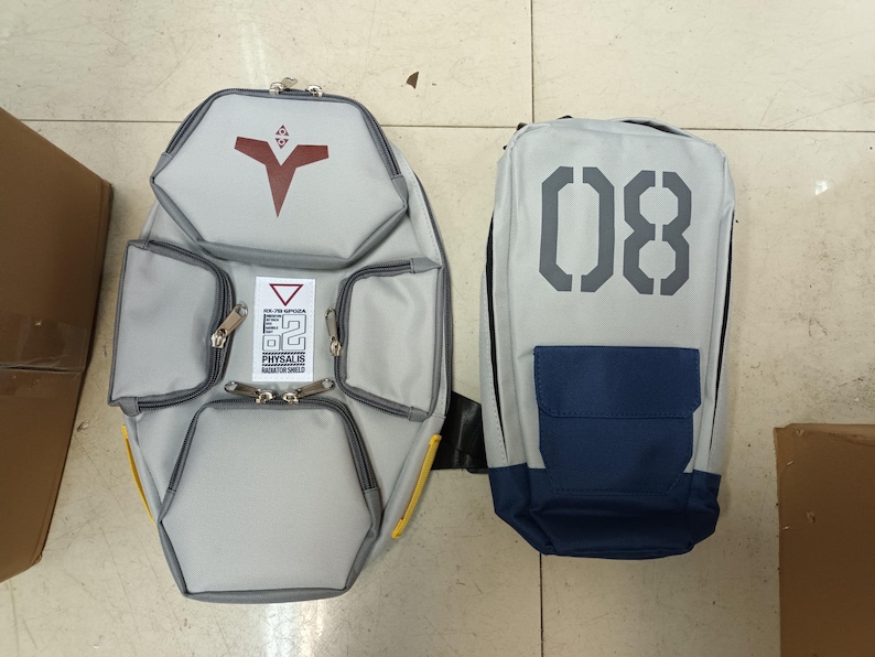 May include: Two gray backpacks. One is a unique shape with a red symbol and multiple pockets. The other is rectangular with the number "08" in gray and a blue pocket. Both have black straps and zippers.