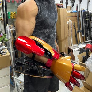 Può includere: Un guanto metallico rosso e oro indossato sull'avambraccio e sulla mano, caratterizzato da un design ad armatura segmentata con finitura lucida. Il pezzo include componenti meccanici visibili, cablaggi interni e cinghie nere di fissaggio.