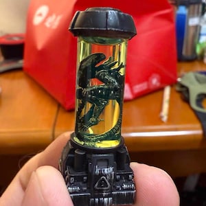Puede incluir: Una figura Alienígena negra y amarilla encerrada en un recipiente cilíndrico. La figura verde oscuro está en un líquido amarillo. El recipiente tiene una tapa y una base negras con un símbolo de peligro.