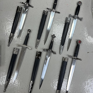 Könnte beinhalten: Sammlung mittelalterlicher Schwerter mit silbernen Klingen und schwarzen Griffen. Jedes Schwert hat ein einzigartiges Griffdesign, einige mit aufwendigen Details. Schwarze Scheiden mit silbernen Akzenten begleiten die Schwerter und bilden eine historische Waffenausstellung.
