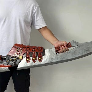 Puede incluir: Una espada grande, de color plateado, con una hoja curva. La empuñadura y la guarda están detalladas con cuero rojo y marrón, detalles metálicos y remaches decorativos. La espada es sostenida por una persona.