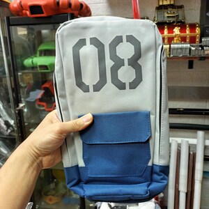 May include: A rectangular gray and blue backpack with the number "08" printed on the front. The backpack has a blue pocket and a black zipper. The bag is held by a person's hand.