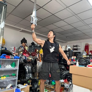 May include: A silver sword with a detailed hilt is held aloft. The sword has a long, straight blade and a decorative handle. The person holding the sword is wearing black shorts and a black vest. The background is filled with various action figures and collectibles.
