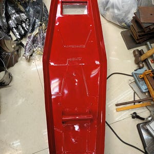 K&ouml;nnte beinhalten: Ein leuchtend rotes, rechteckiges Schild mit einem Sichtfenster und einem Griff. Das Schild hat eine gl&auml;nzende Oberfl&auml;che und scheint aus einem strapazierf&auml;higen Material gefertigt zu sein. Andere R&uuml;stungsteile sind im Hintergrund zu sehen.