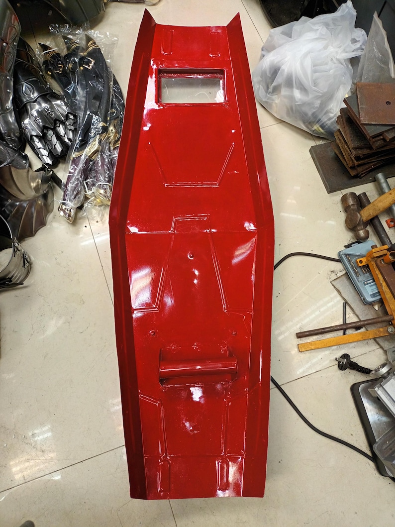 K&ouml;nnte beinhalten: Ein leuchtend rotes, rechteckiges Schild mit einem Sichtfenster und einem Griff. Das Schild hat eine gl&auml;nzende Oberfl&auml;che und scheint aus einem strapazierf&auml;higen Material gefertigt zu sein. Andere R&uuml;stungsteile sind im Hintergrund zu sehen.