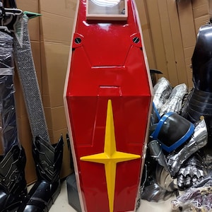 K&ouml;nnte beinhalten: Ein gro&szlig;er, rot-beiger Schild mit gelbem Sternmuster. Der Schild hat ein rechteckiges Sichtfenster und ist wahrscheinlich Teil eines Kost&uuml;ms oder Cosplay-Artikels. Weitere R&uuml;stungsteile sind im Hintergrund zu sehen.