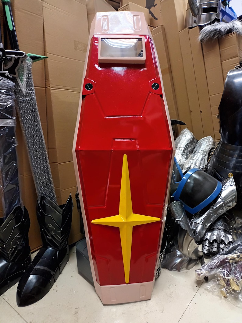 K&ouml;nnte beinhalten: Ein gro&szlig;er, rot-beiger Schild mit gelbem Sternmuster. Der Schild hat ein rechteckiges Sichtfenster und ist wahrscheinlich Teil eines Kost&uuml;ms oder Cosplay-Artikels. Weitere R&uuml;stungsteile sind im Hintergrund zu sehen.