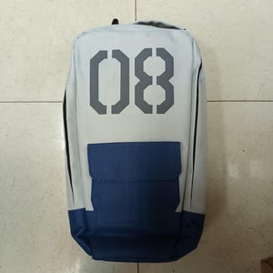 May include: A rectangular backpack with a light gray body, a navy blue pocket, and the number "08" in gray on the front. The backpack has a black handle at the top and black trim along the sides.