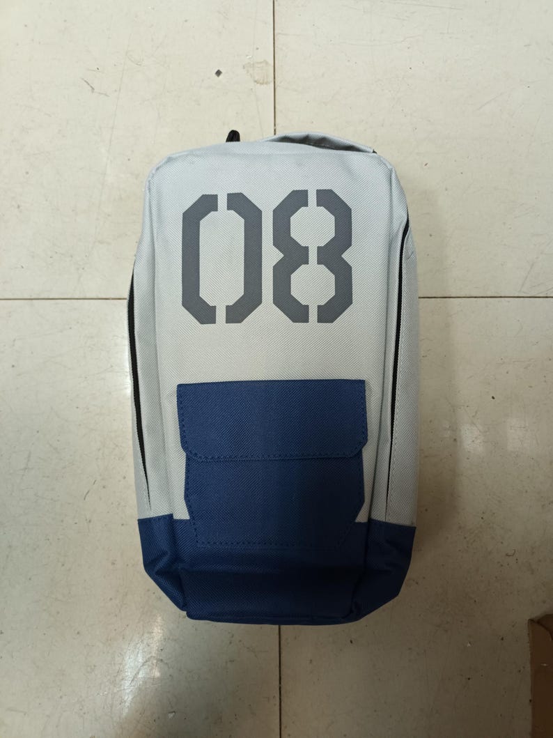 May include: A rectangular backpack with a light gray body, a navy blue pocket, and the number "08" in gray on the front. The backpack has a black handle at the top and black trim along the sides.