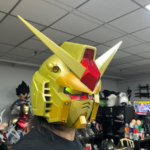 May include: A gold and red Gundam-style helmet with sharp, angular features. The helmet has a metallic finish and includes prominent yellow antennae. The design incorporates red accents and visible internal lighting, suggesting a futuristic aesthetic.