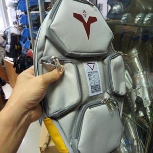 May include: Gray and white shoulder bag with multiple zippered pockets. The bag features a red graphic design and the text "RX-78 GPO2A PHYASALIS RADIATOR SHIELD". A yellow strap is visible on the side.