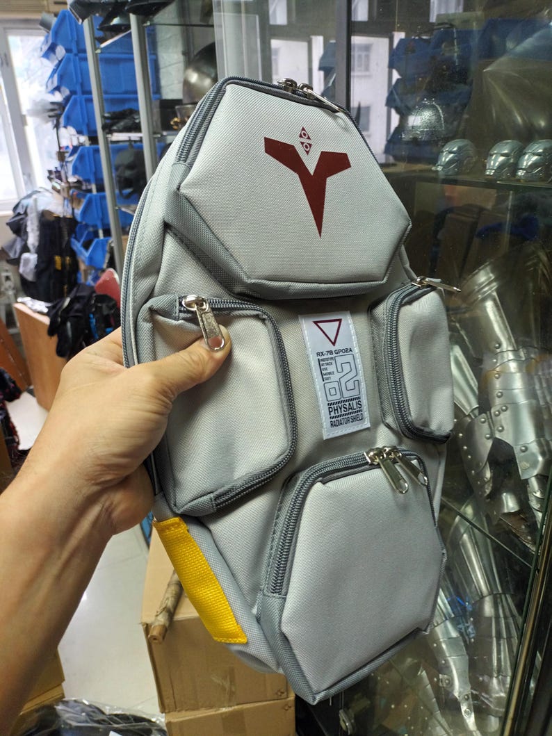 May include: Gray and white shoulder bag with multiple zippered pockets. The bag features a red graphic design and the text "RX-78 GPO2A PHYASALIS RADIATOR SHIELD". A yellow strap is visible on the side.