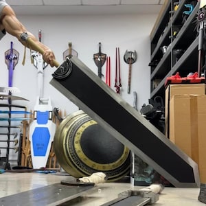May include: A large, heavy-duty sword with a wide, rectangular black blade and a metallic edge. The hilt is wrapped in light-colored material, and the pommel features a circular design. The sword is held at an angle, resting near a round shield.