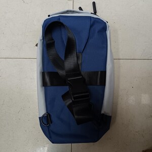 May include: A blue and gray sling bag with a black adjustable strap and buckle. The bag has a D-ring attachment and a textured fabric. The design is simple and modern, suitable for everyday use.