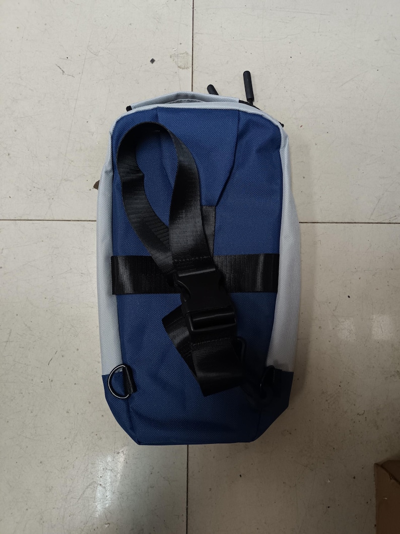 May include: A blue and gray sling bag with a black adjustable strap and buckle. The bag has a D-ring attachment and a textured fabric. The design is simple and modern, suitable for everyday use.