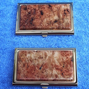 May include: Two gold-toned metal business card holders with a wood-grain pattern. The wood-grain pattern is a reddish-brown color with dark brown streaks.