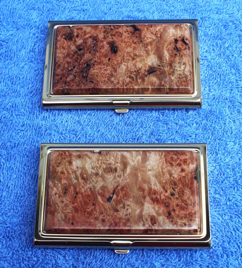 May include: Two gold-toned metal business card holders with a wood-grain pattern. The wood-grain pattern is a reddish-brown color with dark brown streaks.