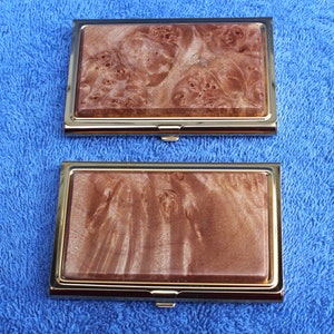 May include: Two gold-trimmed business card holders with a wood grain pattern. The top holder has a light brown wood grain pattern with darker brown swirls. The bottom holder has a darker brown wood grain pattern with lighter brown swirls.