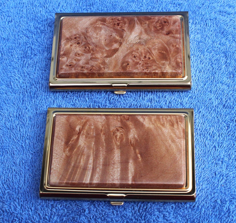 May include: Two gold-trimmed business card holders with a wood grain pattern. The top holder has a light brown wood grain pattern with darker brown swirls. The bottom holder has a darker brown wood grain pattern with lighter brown swirls.