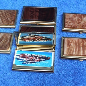 May include: Nine wooden business card holders with gold trim. The holders are made of different types of wood with unique grain patterns. One holder is open, revealing a business card with the text "J.M.'s Workshop, Woodturning and More".