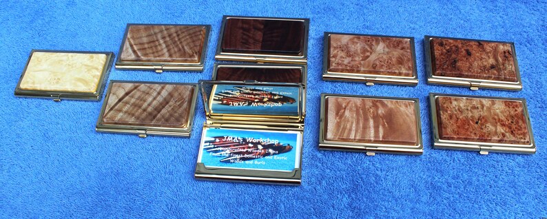 May include: Nine wooden business card holders with gold trim. The holders are made of different types of wood with unique grain patterns. One holder is open, revealing a business card with the text "J.M.'s Workshop, Woodturning and More".