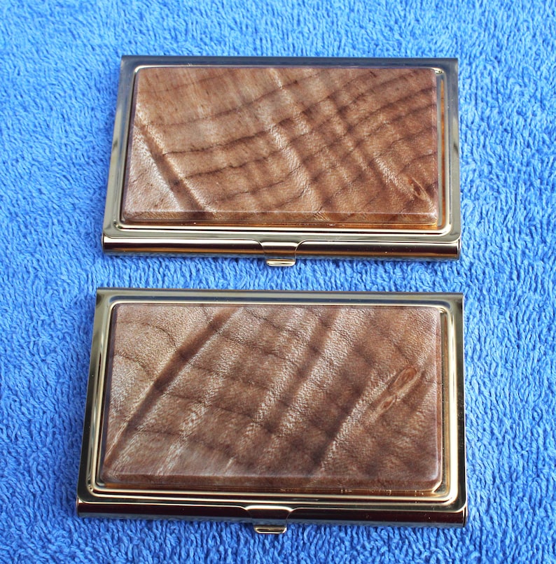 May include: Two gold-trimmed business card holders with a wood grain pattern. The wood grain is a light brown with darker brown streaks.