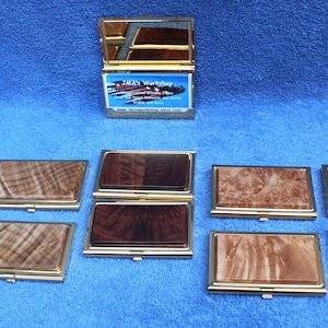 May include: Ten different wooden business card holders with gold trim. The holders are arranged in two rows of five. The holders are made of different types of wood, including maple, walnut, and cherry. The holders are all closed and have a gold hinge.