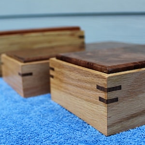 May include: Four wooden boxes of varying sizes, with intricate joinery details. The largest box has a dark wood top and light wood sides, while the smaller boxes are all light wood. The boxes are arranged in a row on a blue towel.