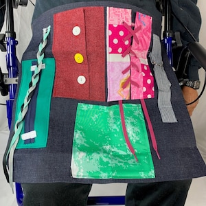 May include: A sensory activity mat with various textures and colors. It features a red fabric square with three buttons, a green fabric square with a zipper, a pink fabric square with a lace-up design, and a green fabric square with a marble pattern.