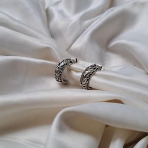 May include: Two silver hoop earrings with an elaborate, swirling pattern. The earrings are displayed on white satin fabric, which accentuates their detailed design. The earrings have a shiny finish, reflecting light.
