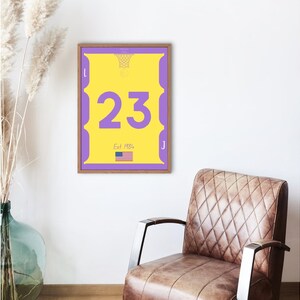 Puede incluir: Un estampado enmarcado con un diseño de camiseta de baloncesto amarilla con el número 23 en morado. El estampado incluye un gráfico de red de baloncesto, el texto "Est 1984" y una pequeña bandera estadounidense. El marco es marrón.