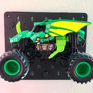 May include: A green and yellow toy monster truck with large black tires and green rims. The truck has a dragon design on the body and is mounted on a black pegboard. The text "DRAGONOID" is visible on the truck.