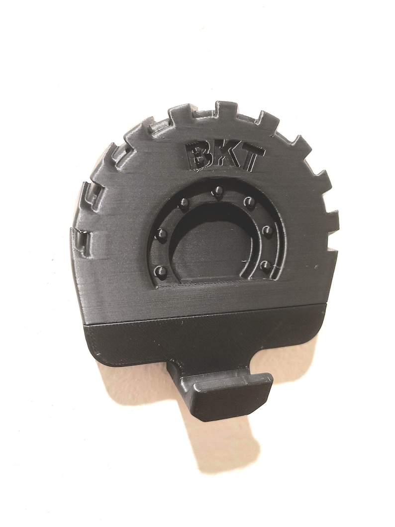May include: A black, 3D-printed tire-shaped object with the letters "BKT" embossed on the tire. The tire has a gear-like outer edge and a central circular design with rivets. A rectangular hook extends from the bottom.