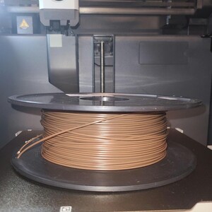 May include: A brown filament spool sits on a 3D printer bed. The spool is filled with tightly wound brown filament. The printer bed has the dimensions 256 x 256 x 256 mm.