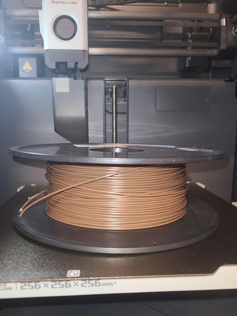 May include: A brown filament spool sits on a 3D printer bed. The spool is filled with tightly wound brown filament. The printer bed has the dimensions 256 x 256 x 256 mm.