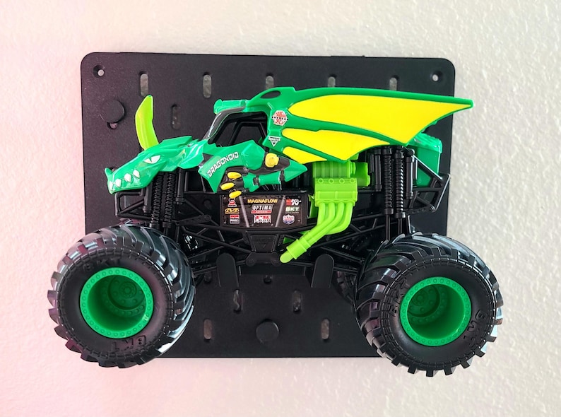 May include: A green and yellow toy monster truck with large black tires and green rims. The truck has a dragon design on the body and is mounted on a black pegboard. The text "DRAGONOID" is visible on the truck.