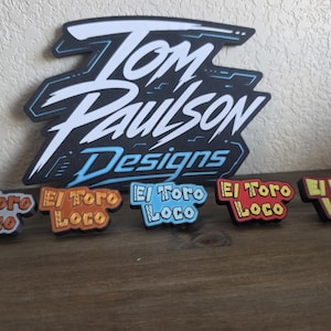 Pode incluir: Uma placa preta grande com texto branco e azul escrito Tom Paulson Designs, posicionada atrás de cinco placas retangulares menores e coloridas. Cada placa pequena apresenta o texto El Toro Loco em cores distintas: cinza, laranja, azul, vermelho e amarelo.
