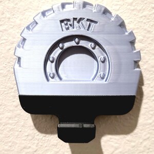 May include: A light gray, 3D-printed object with the letters "BKT" on top. The object has a circular design with a black base and a mounting tab. The design resembles a tire tread.