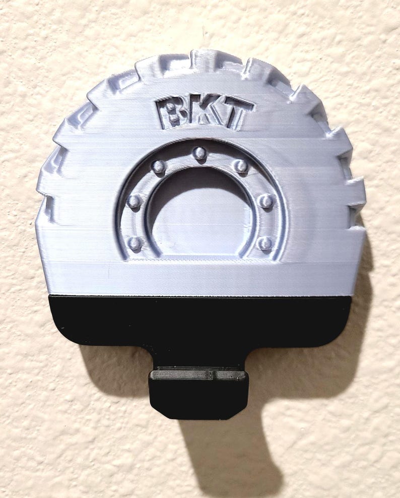 May include: A light gray, 3D-printed object with the letters "BKT" on top. The object has a circular design with a black base and a mounting tab. The design resembles a tire tread.