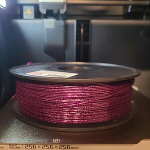 May include: A spool of magenta 3D printer filament sits on a printer bed. The filament is tightly wound and appears to have a glittery texture. The printer bed has the dimensions 256 x 256 x 256mm.