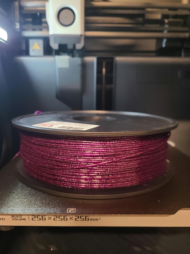 May include: A spool of magenta 3D printer filament sits on a printer bed. The filament is tightly wound and appears to have a glittery texture. The printer bed has the dimensions 256 x 256 x 256mm.