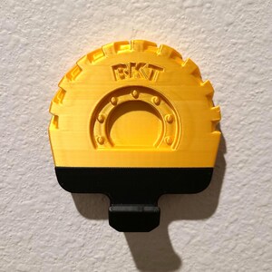 May include: A yellow and black 3D-printed object with the letters "BKT" on the top. The object has a semi-circular shape with a gear-like edge and a black handle. The design includes a circular pattern with rivets.