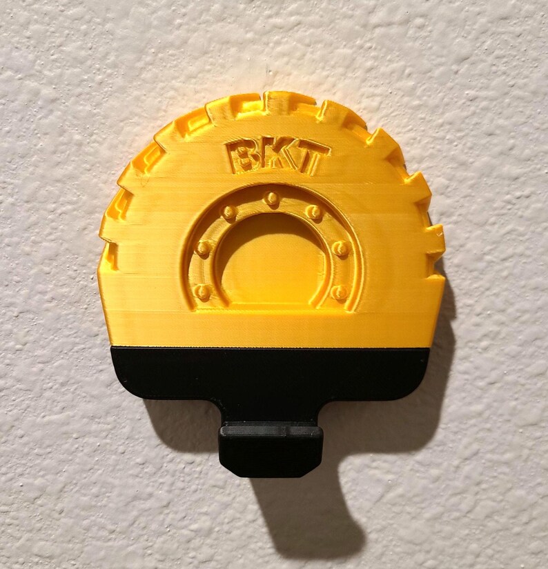 May include: A yellow and black 3D-printed object with the letters "BKT" on the top. The object has a semi-circular shape with a gear-like edge and a black handle. The design includes a circular pattern with rivets.