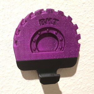 May include: A purple, glittery, gear-shaped object with the letters "BKT" embossed on the top. The object has a black base with a small, rectangular protrusion. The object is likely a decorative or functional item.